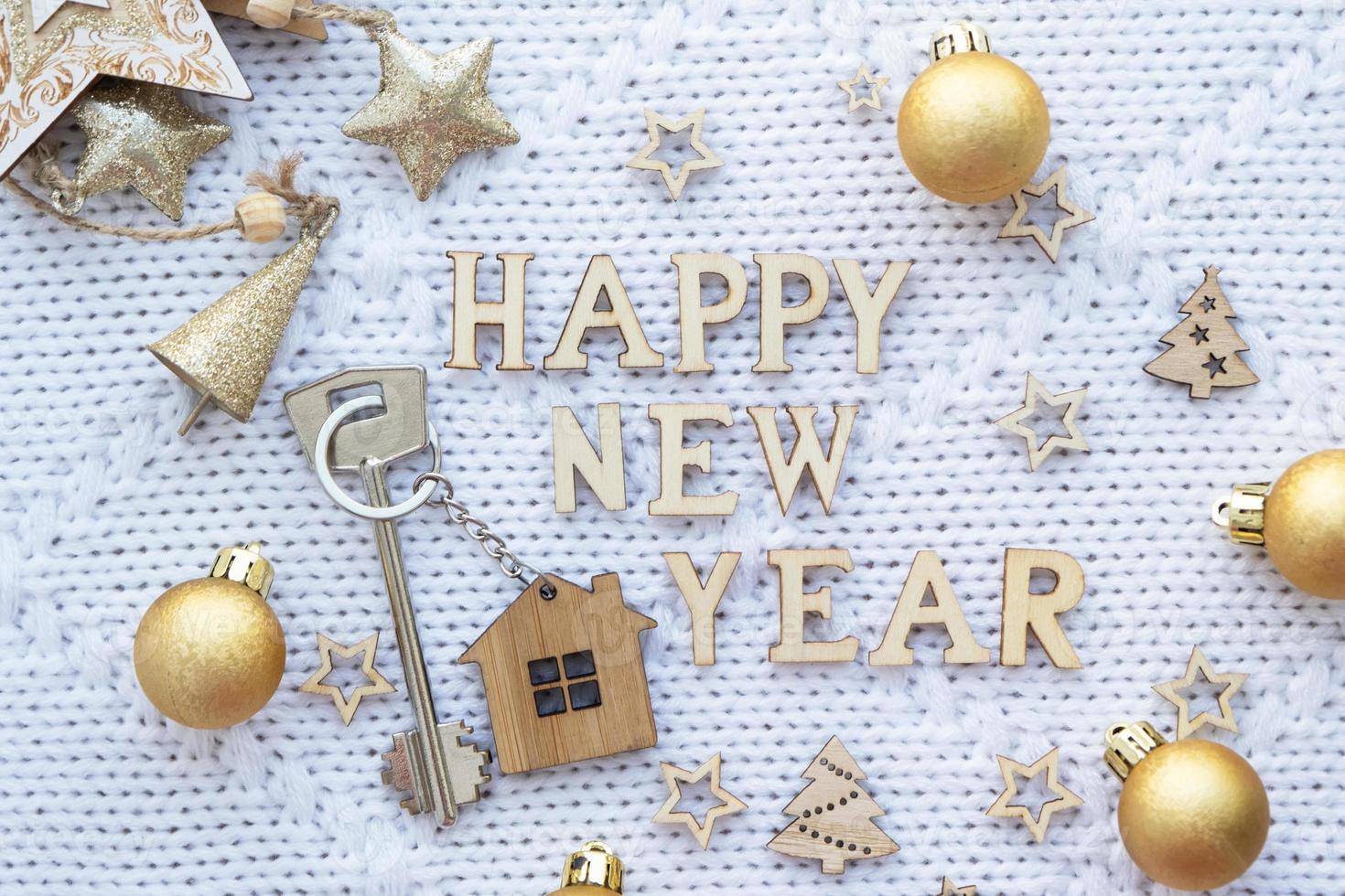 House key with keychain cottage on a festive knitted background with sequins, stars, lights of garlands. Happy New Year-wooden letters, greeting card. Purchase, construction, relocation, mortgage photo