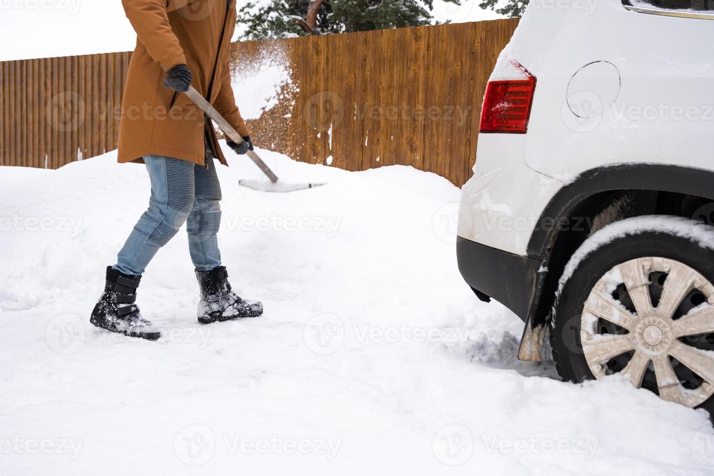 A man in winter cleans snow with a shovel in the yard of a house in the parking lot. Snowfall, difficult weather conditions, the car is stalling, digging up the passage photo