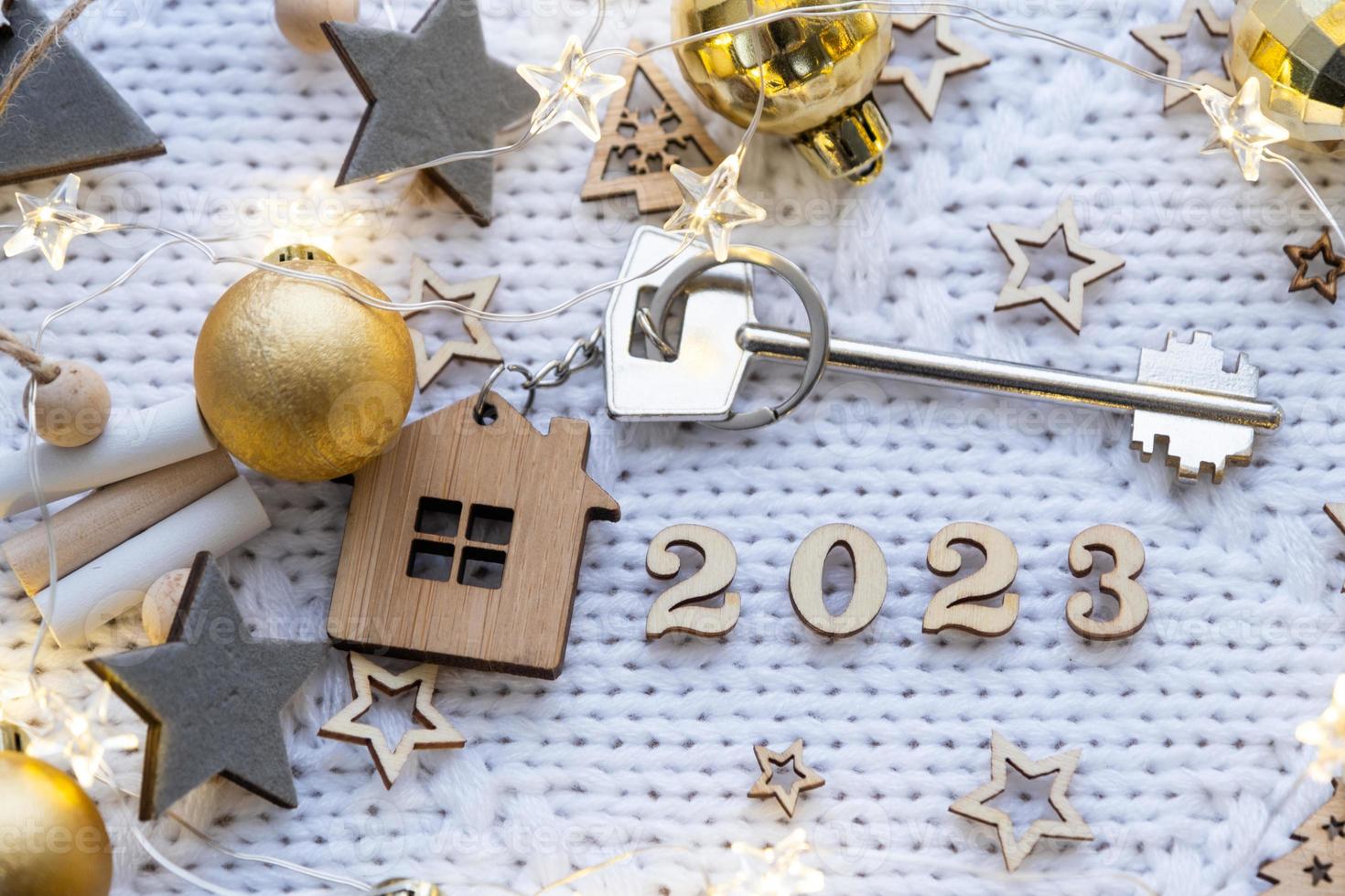 House key with keychain cottage on festive knitted background with stars, lights of garlands. Happy New Year 2023-wooden letters, greeting card. Purchase, construction, relocation, mortgage photo