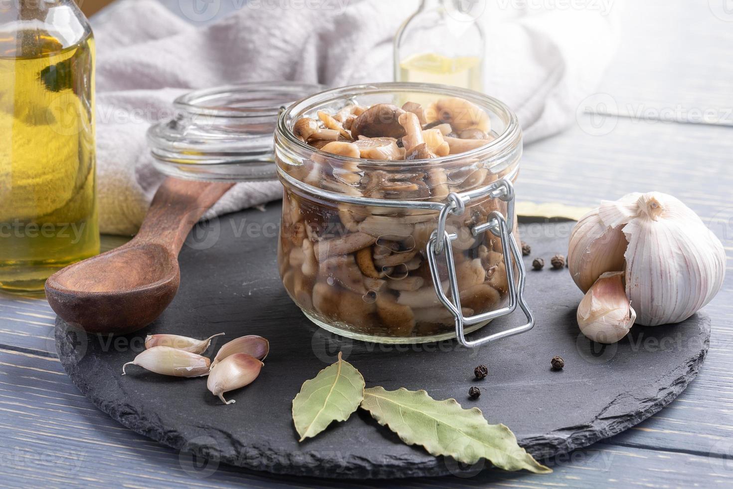 Homemade pickled honey agarics in open glass jar, spices, spoon, napkin, oil bottles on grey wooden table. Close-up. photo