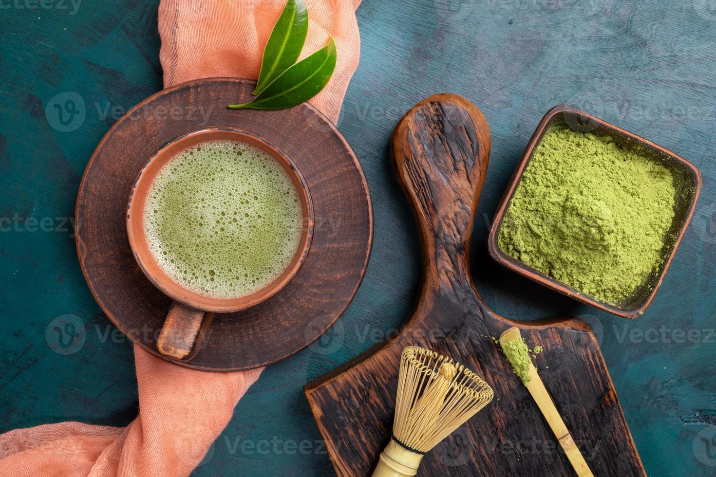 Green matcha tea and powder in brown ceramic cups and whisk and spoon on wooden serving board on emerald backdrop. photo