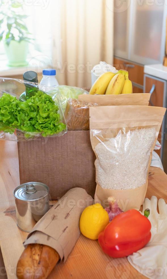 Fresh food products in cardboard box and on wooden table in kitchen interior. Safe delivery. photo