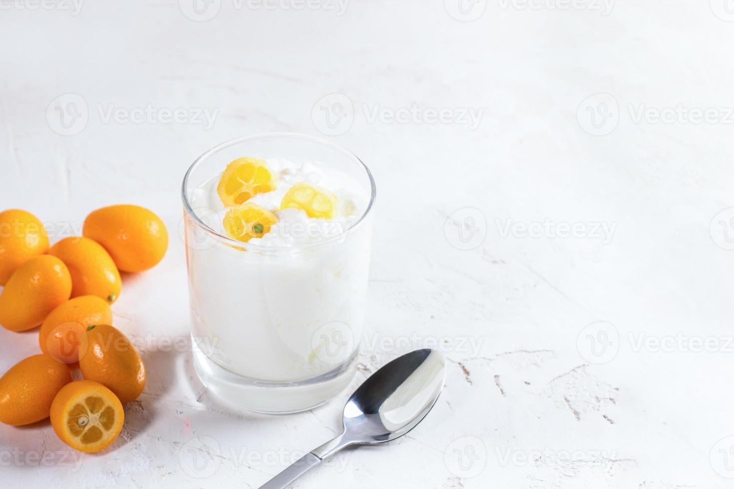 yogur blanco con rodajas de kumquat en vidrio, frutas enteras y cuchara metálica sobre fondo blanco. foto