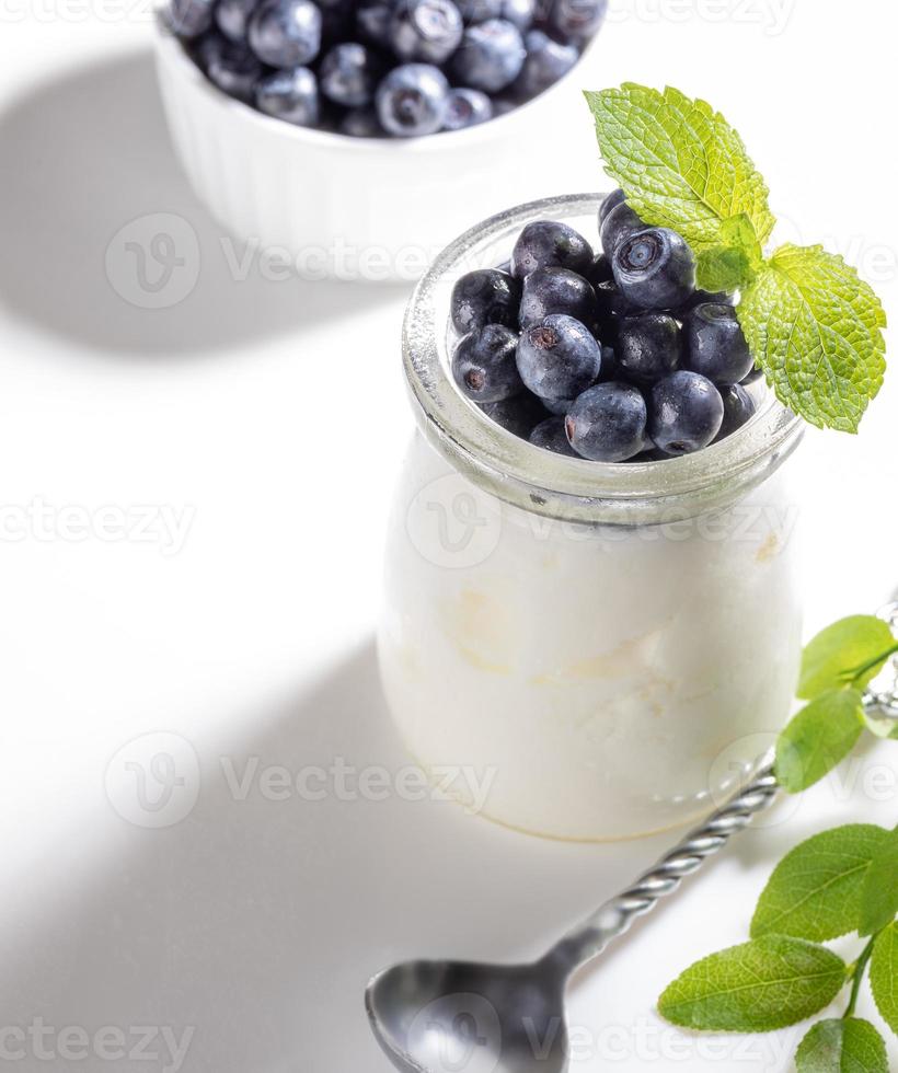 Natural yogurt in glass jar with fresh forest blueberries on white background. Copy space. photo