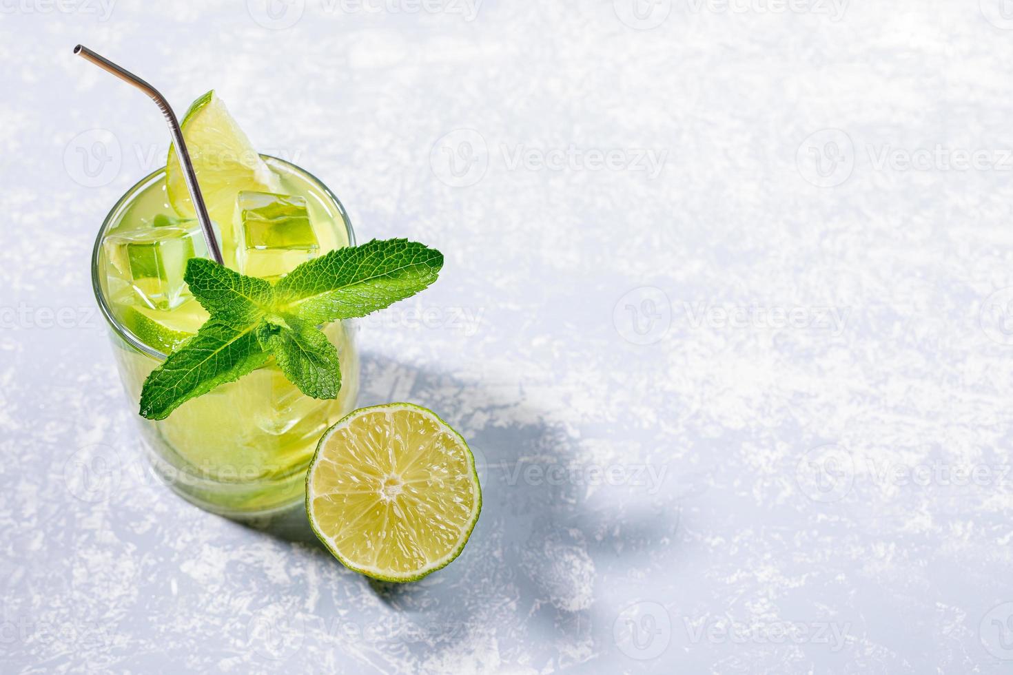 One glass with iced green matcha tea with lime, ice, fresh mint and metal drinking straw on light grey with shadows. photo