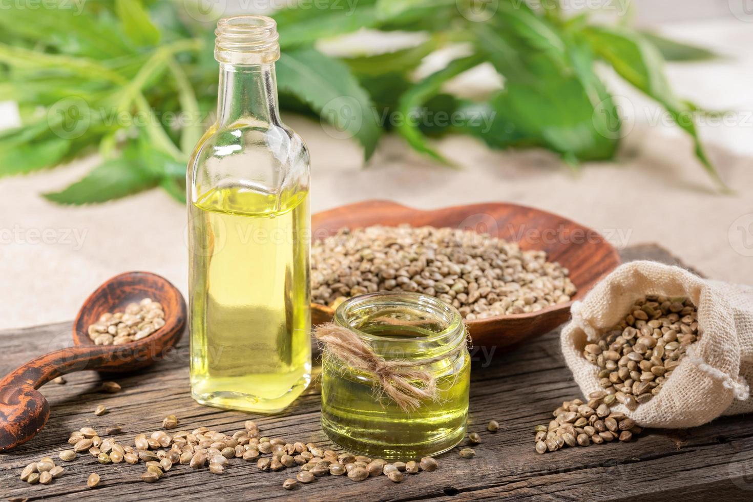 Hemp oil in glass jar and bottle, seeds in ceramic plate and spoon and in sack on wooden board, with leaves on backdrop. photo