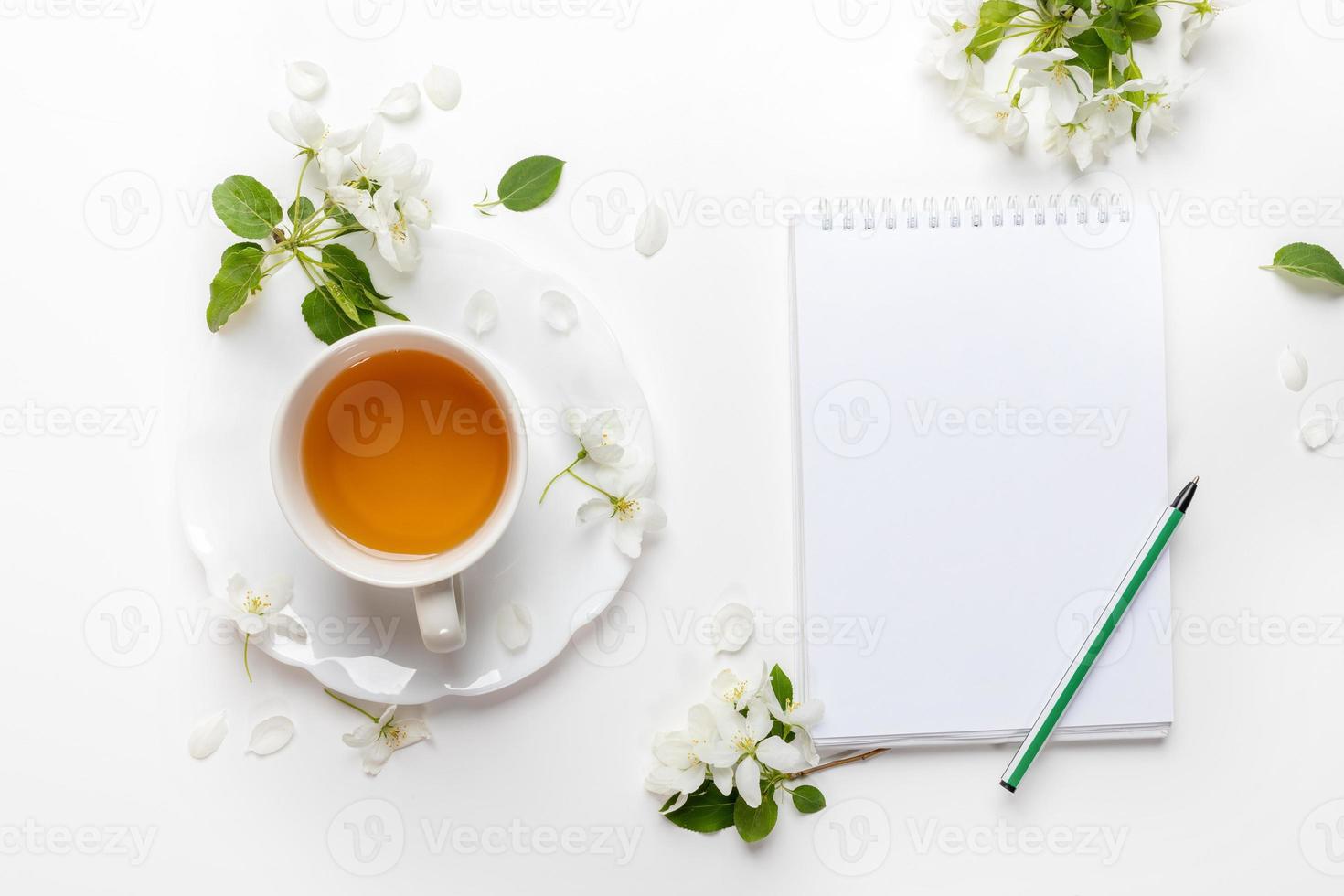 White cup of tea, empty notebook with pen, white apple tree flowers on white. Tea time. Top view. photo