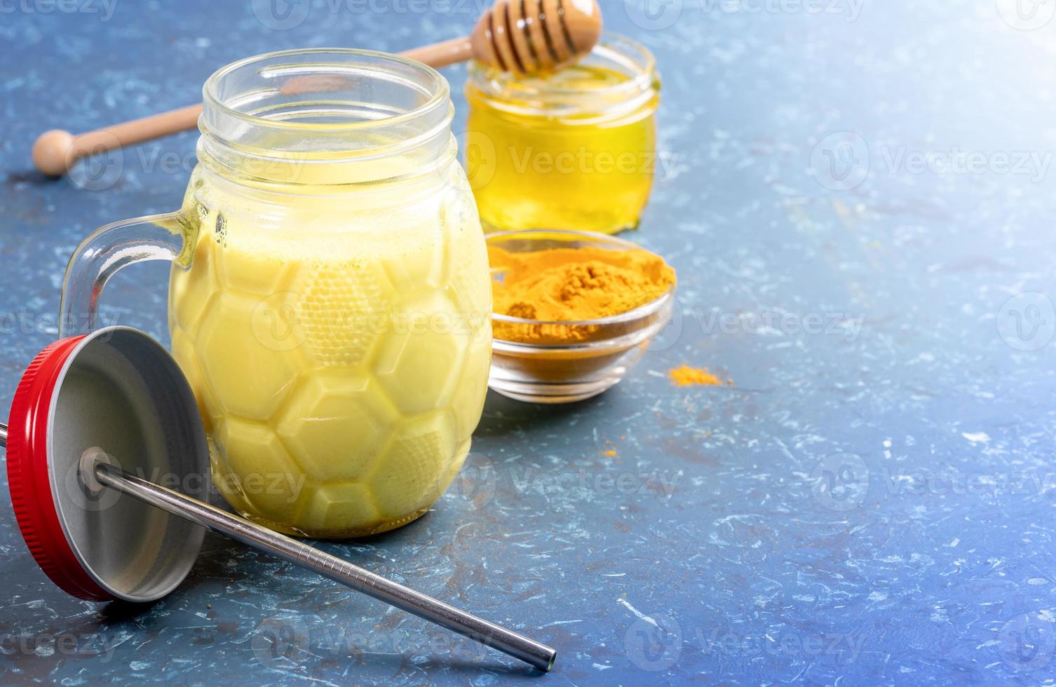 Mason jar of golden turmeric milk with metal straw, curcuma powder and honey jar on blue. Selective focus, copy space. photo