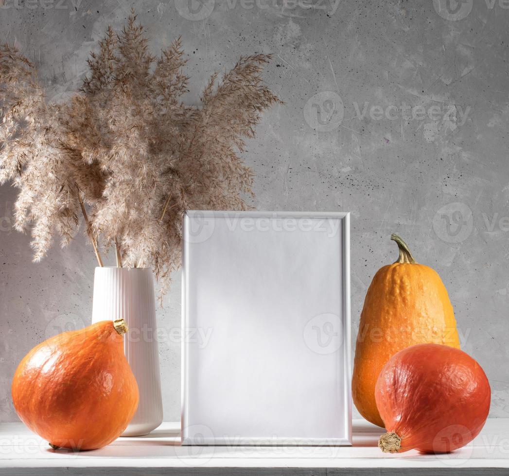 Festive fall mock up with pumpkins, empty frame, dry grass on white table on concrete wall backdrop. photo