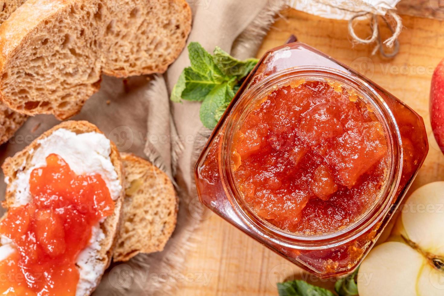 Close-up open glass jar  full of apple jam, sliced  bread, smeared with curd and apple jam on wooden table. photo