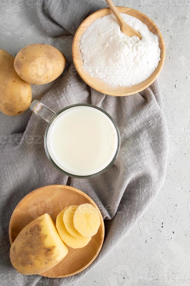 leche en polvo de patata en taza, plato de madera con polvo de patata, tubérculos en servilleta gris sobre hormigón. foto