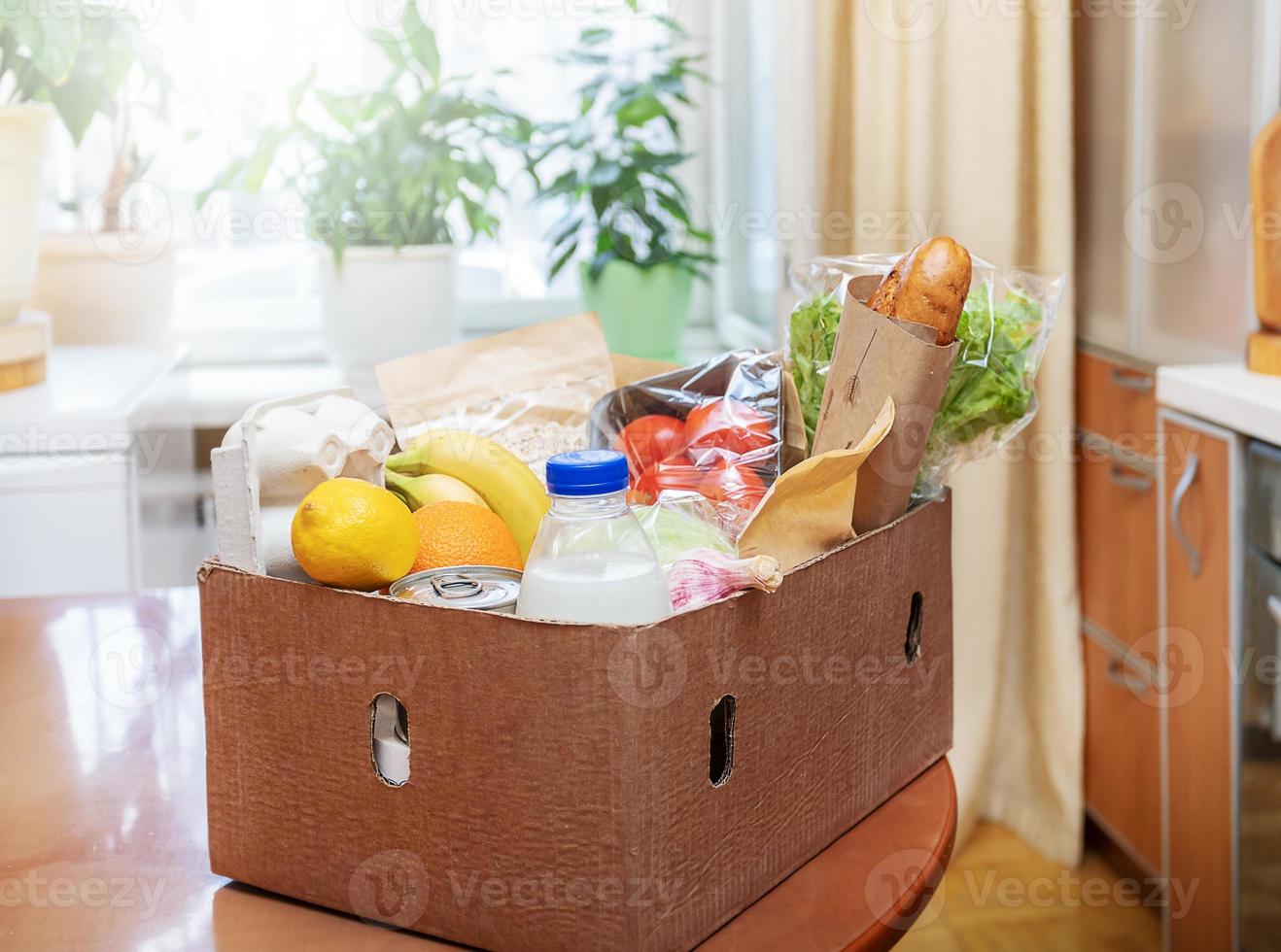 Cardboard box with food products on wooden table in kitchen interior against window with houseplants. Safe delivery. photo