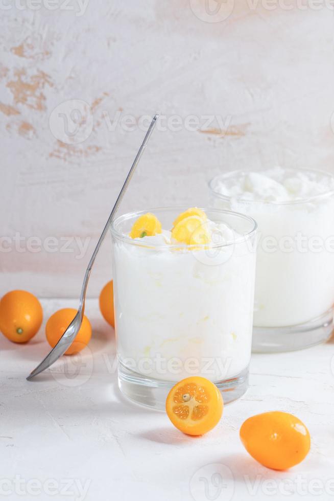 Two glasses with  yogurt, one standing spoon, whole and cut kumquats on white table on light background. photo