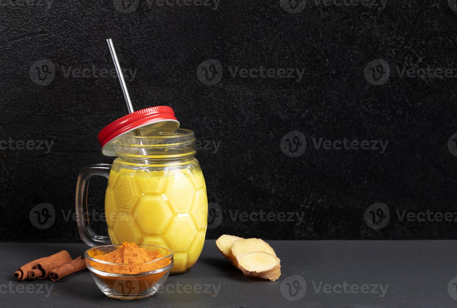 Mason jar of golden turmeric milk with metal straw, ingredients for its cooking on black background. Copy space. photo