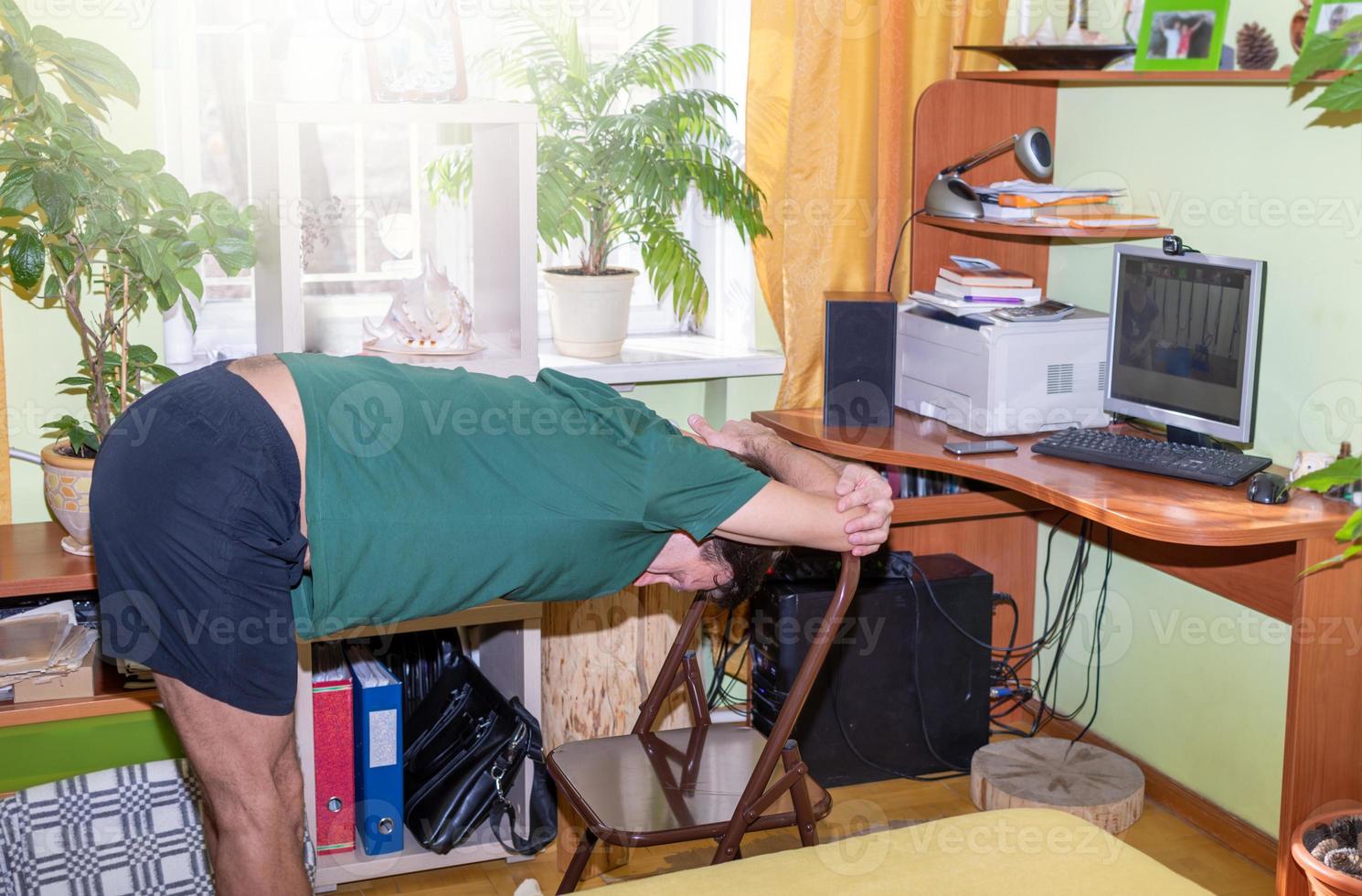 Mature man doing yoga exercise - tilt to chair with before computer monitor in sunny room at home. Remote sport. photo