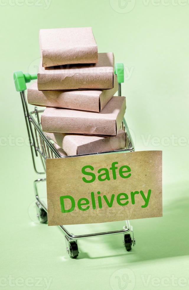Stack of packed boxes in mini shopping trolley with label Safe Delivery. Light green backdrop. Vertical orientation. photo