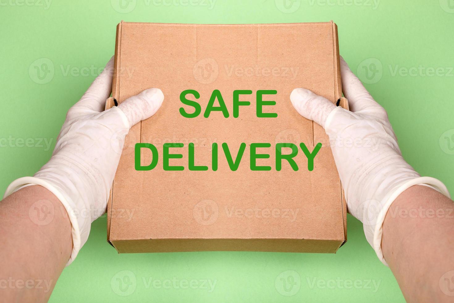 Female hands in white latex gloves are holding out brown cardboard box with words Safe Delivery on green backdrop. photo