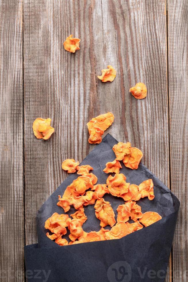 Orange healthy carrot chips falling from black paper bag to old wooden table. photo