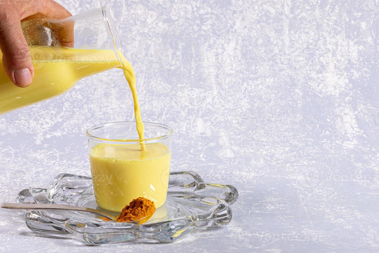 Organic turmeric milk for immunity. Female hand pours golden milk from bottle into glass on light grey background. photo