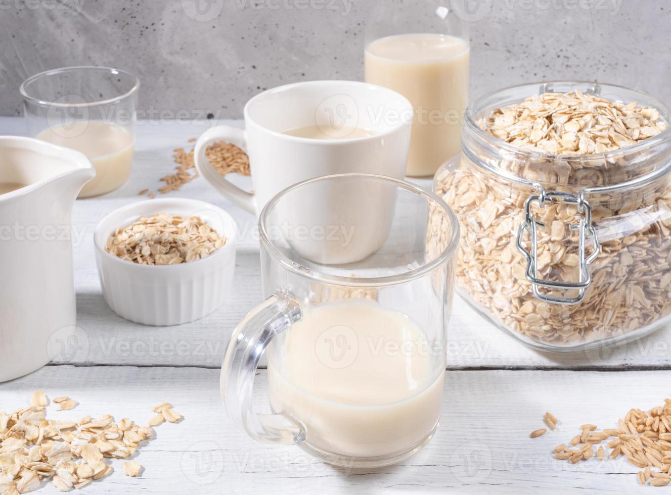 Close-up set of glasses and bottle with oat milk, jar with oat flakes on white wooden table near concrete wall. photo