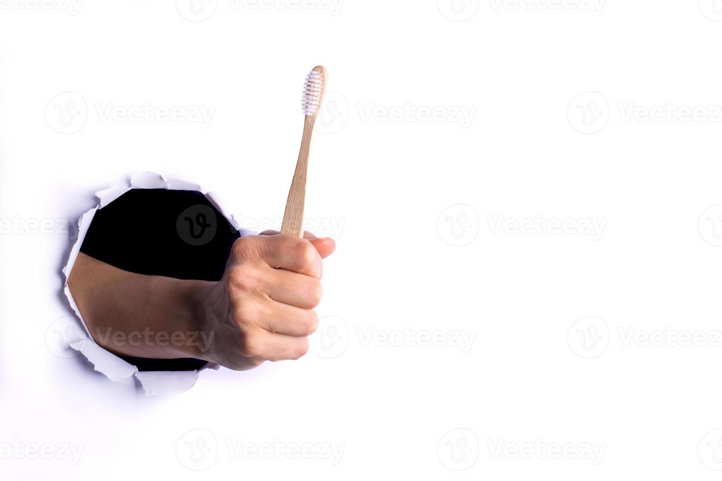 Female hand holding one wooden bamboo toothbrush through torn hole in white paper wall. Zero waste and no plastic. photo