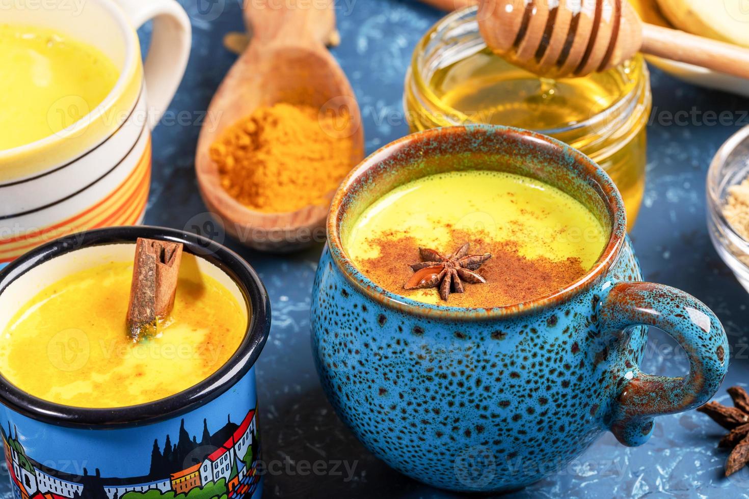 Different cups with golden turmeric milk, curcuma powder and honey on blue textured backdrop. Close-up, selective focus. photo