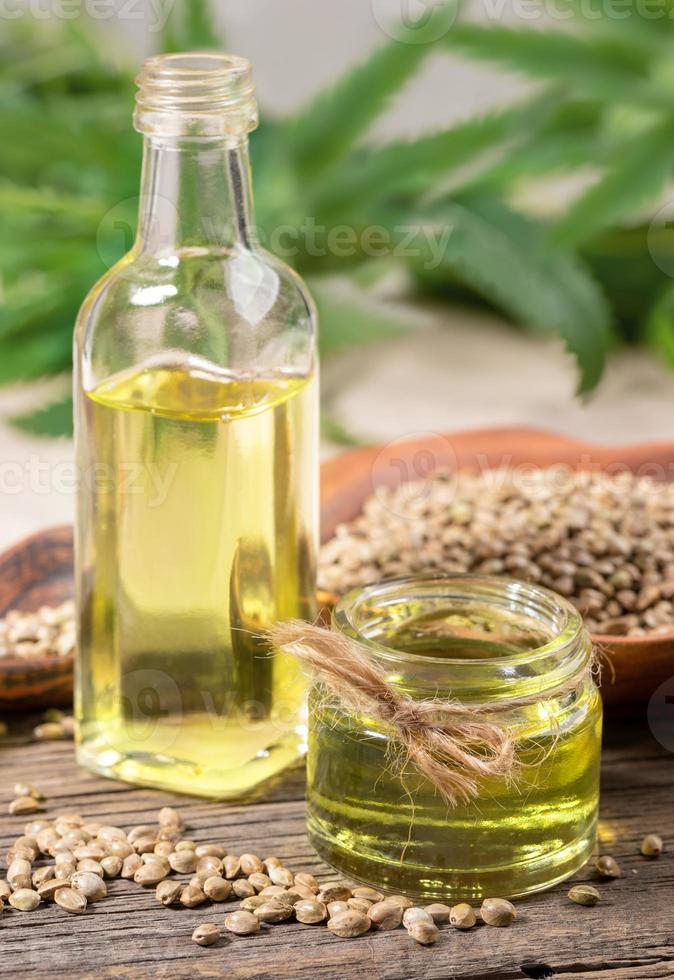 Hemp oil in glass jar and bottle, cannabis seeds in brown ceramic plate and on wooden board and leaves on backdrop. photo