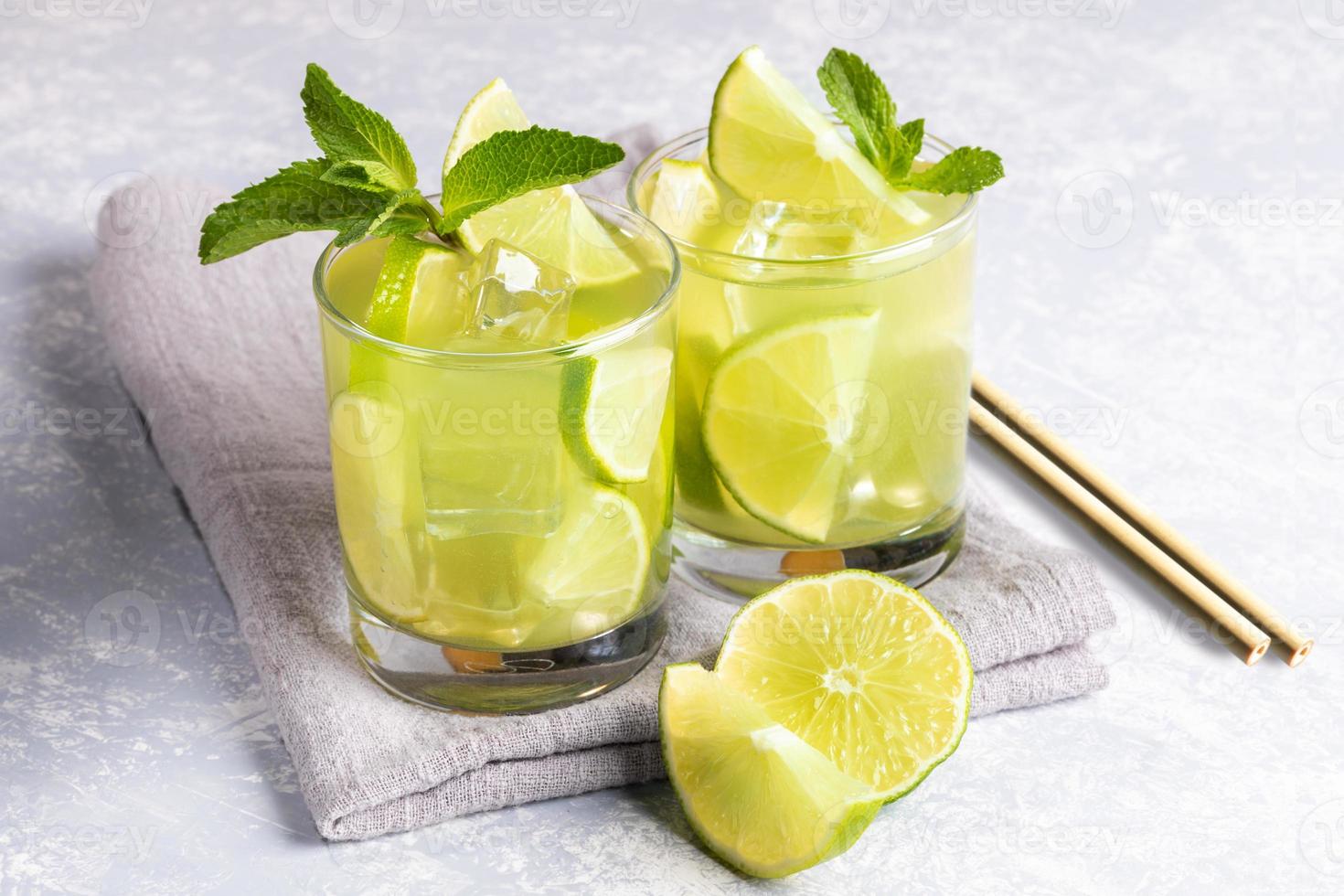 Two glasses of iced green matcha tea with lime, ice, fresh mint, bamboo drinking straws on grey napkin on light grey. photo