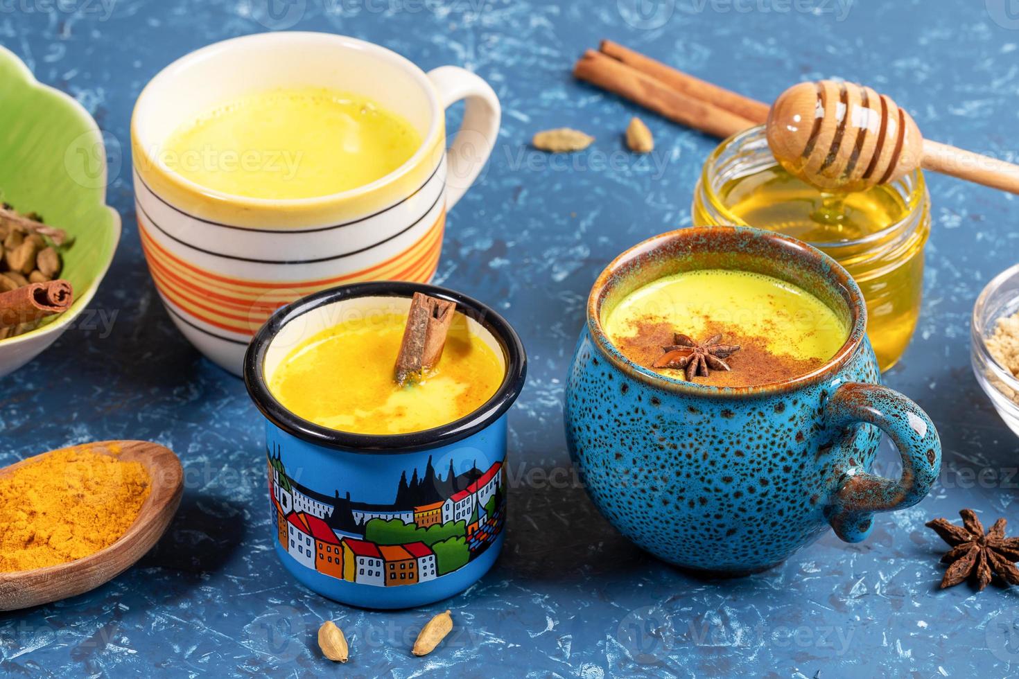 Different cups with golden turmeric milk and ingredients for its cooking on blue backdrop. Close-up, selective focus. photo