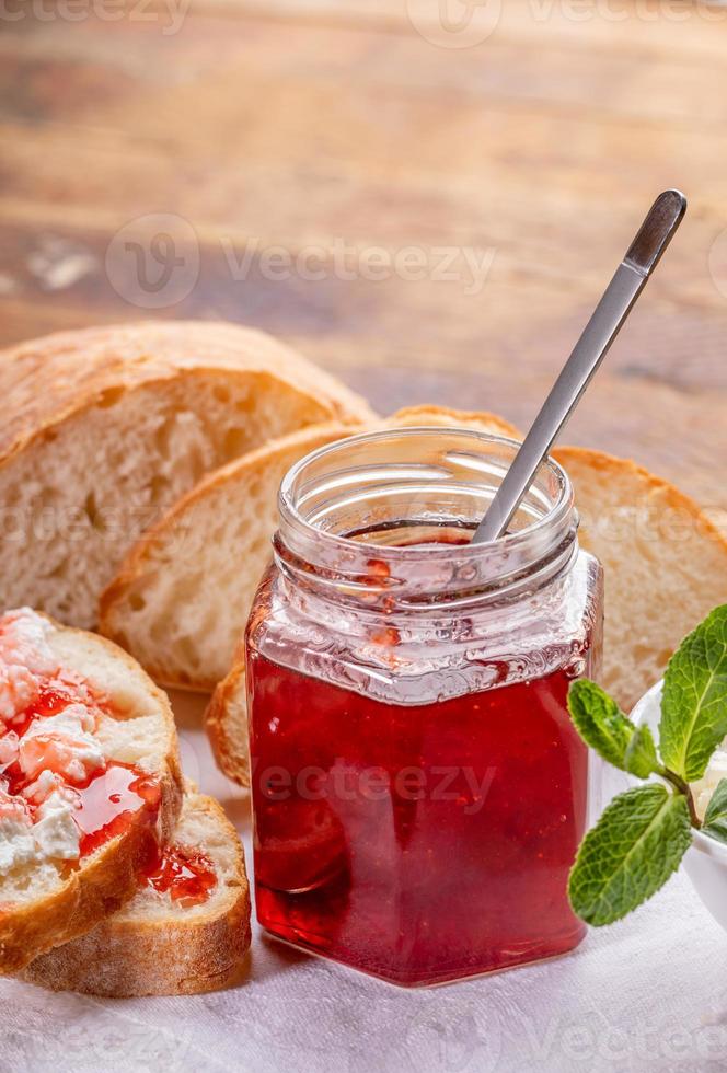 tarro de cristal con mermelada de fresa, pan, tostadas con cuajada y mermelada en servilleta blanca sobre mesa de madera. foto