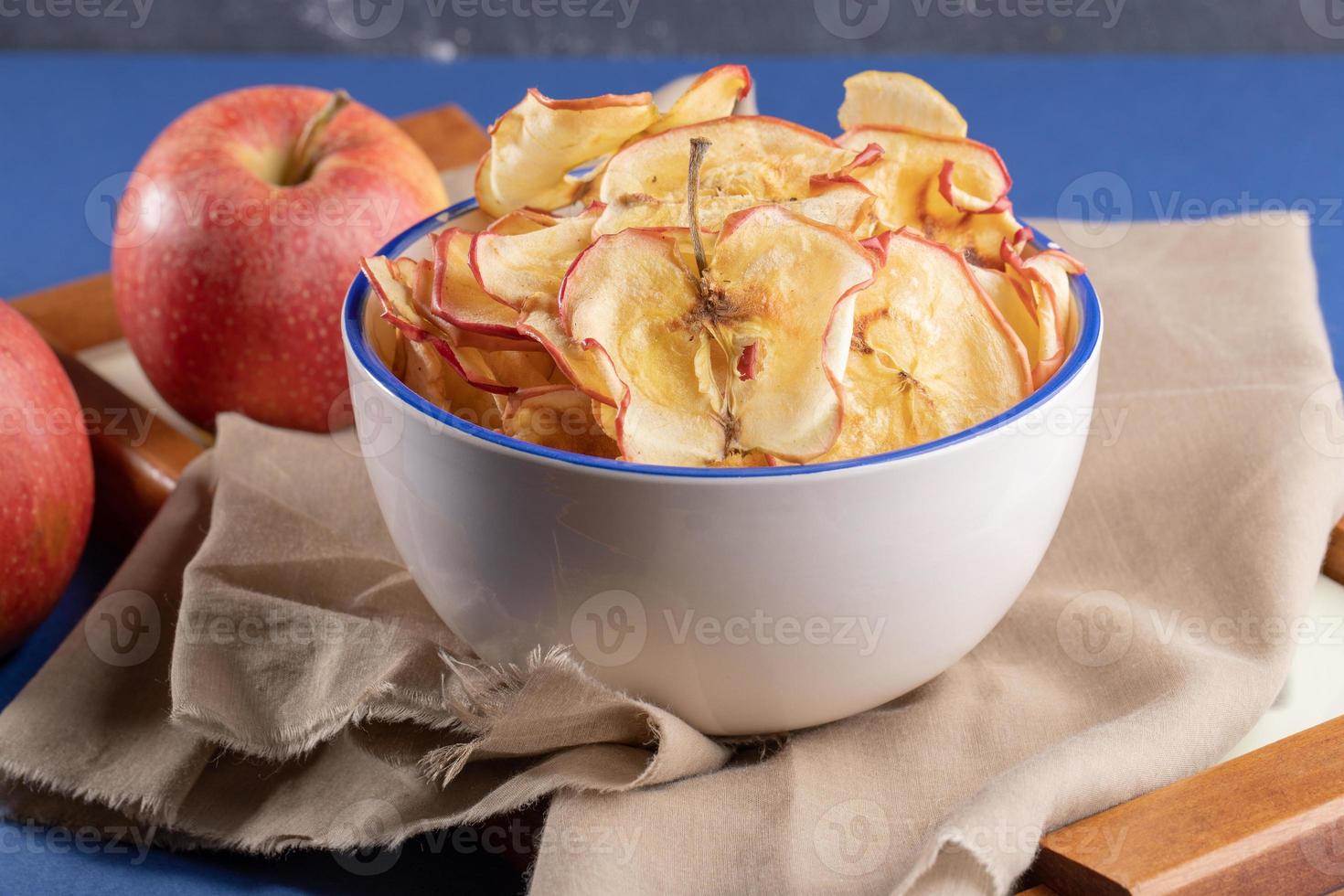 chips de manzana de primer plano en taza y manzanas frescas sobre tela beige sobre bandeja de madera sobre fondo azul. foto