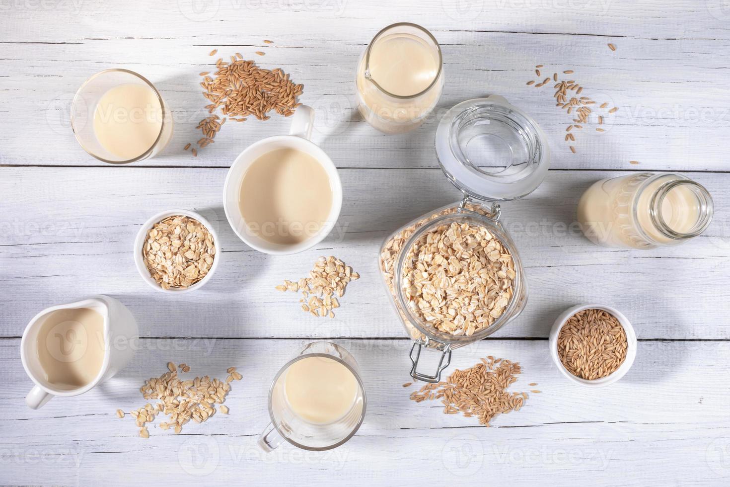 conjunto de diferentes recipientes de vidrio con leche de avena, semillas de avena y copos sobre una mesa de madera blanca. foto