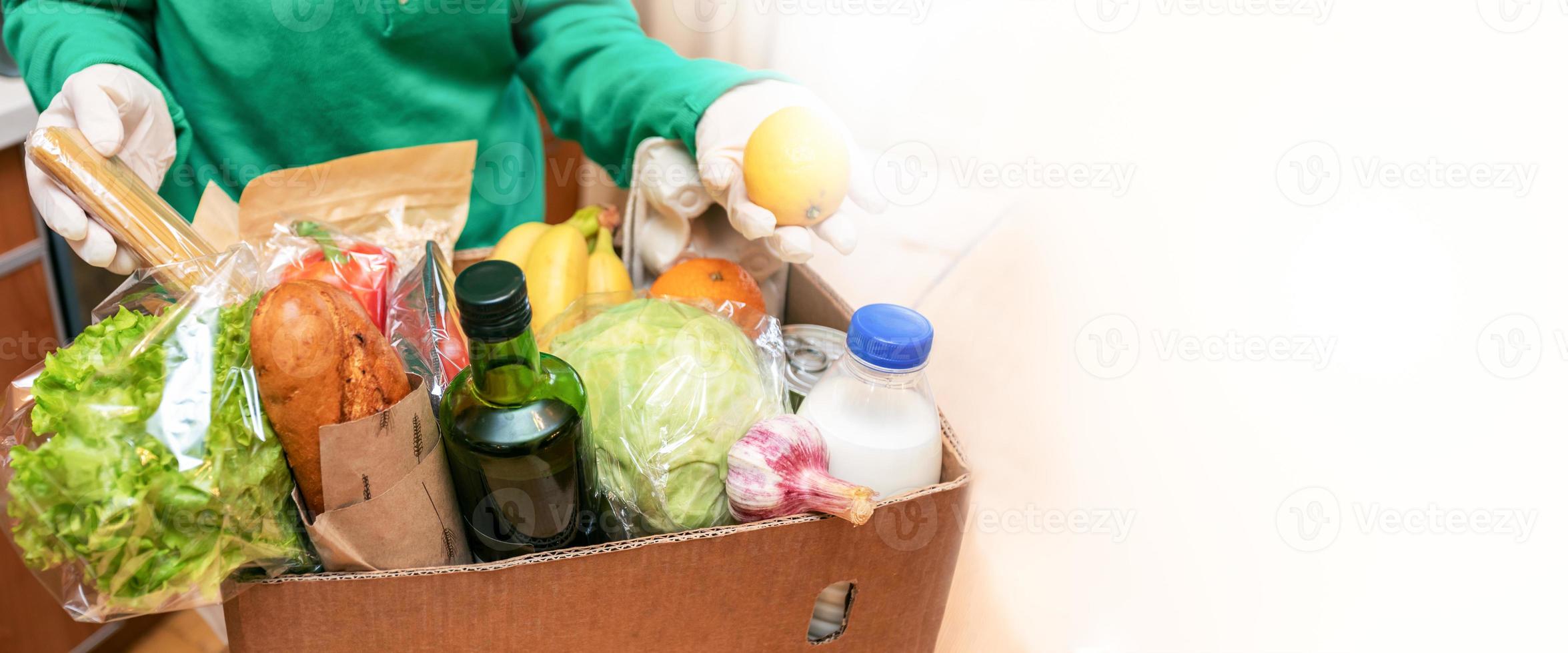 Woman in protective gloves taking food products out of delivered box in kitchen. Safe delivery. Banner, copy space. photo