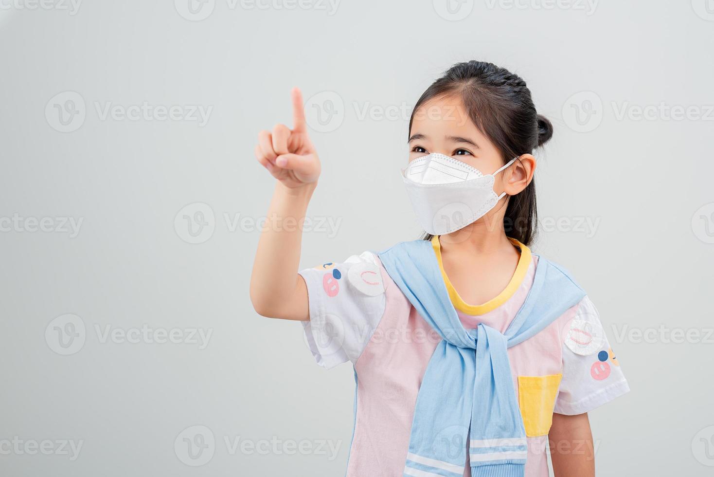 niña asiática con mascarilla respiratoria para proteger el brote de coronavirus y apuntando con la mano al fondo en blanco, nuevo virus covid-19 foto