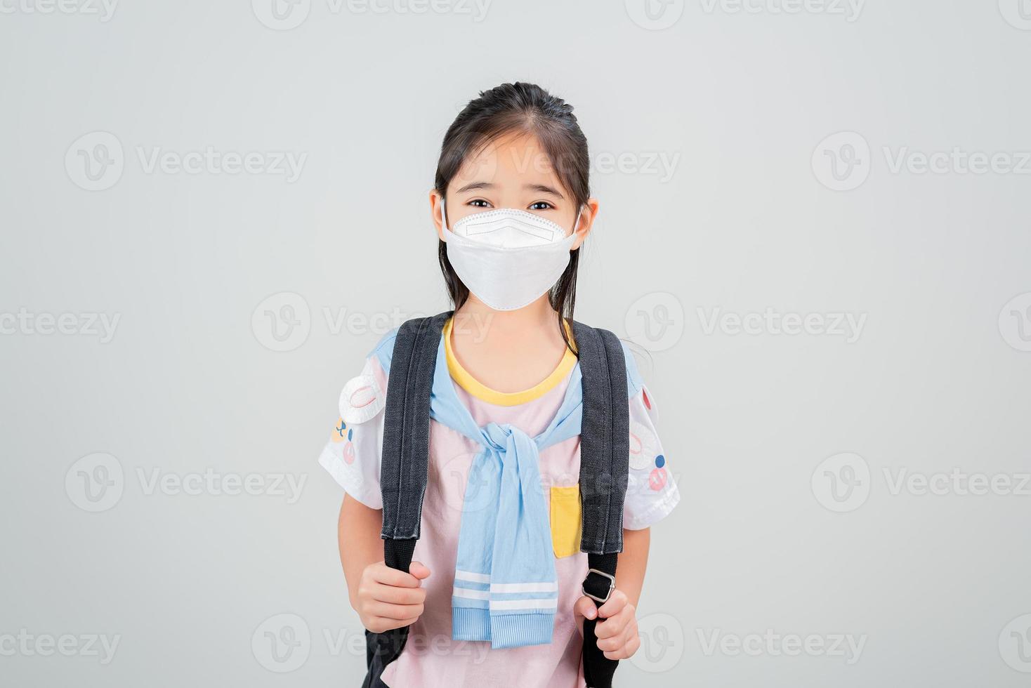 Asian little child girl carry a backpack wearing respirator mask to protect coronavirus outbreak and pointing hand to blank background, New virus Covid-19 photo