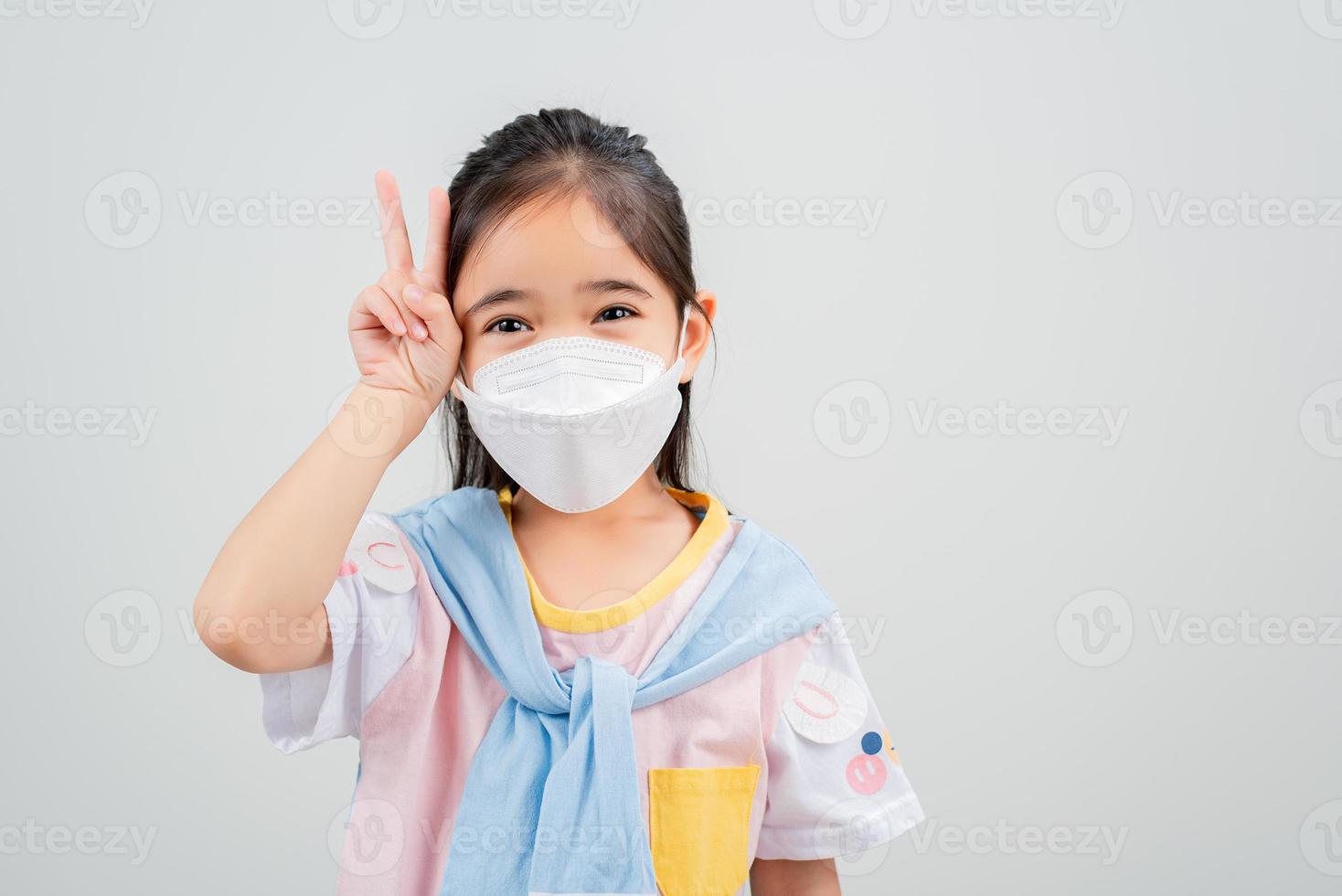 Asian little child girl wearing respirator mask to protect coronavirus outbreak and pointing hand to blank background, New virus Covid-19 photo