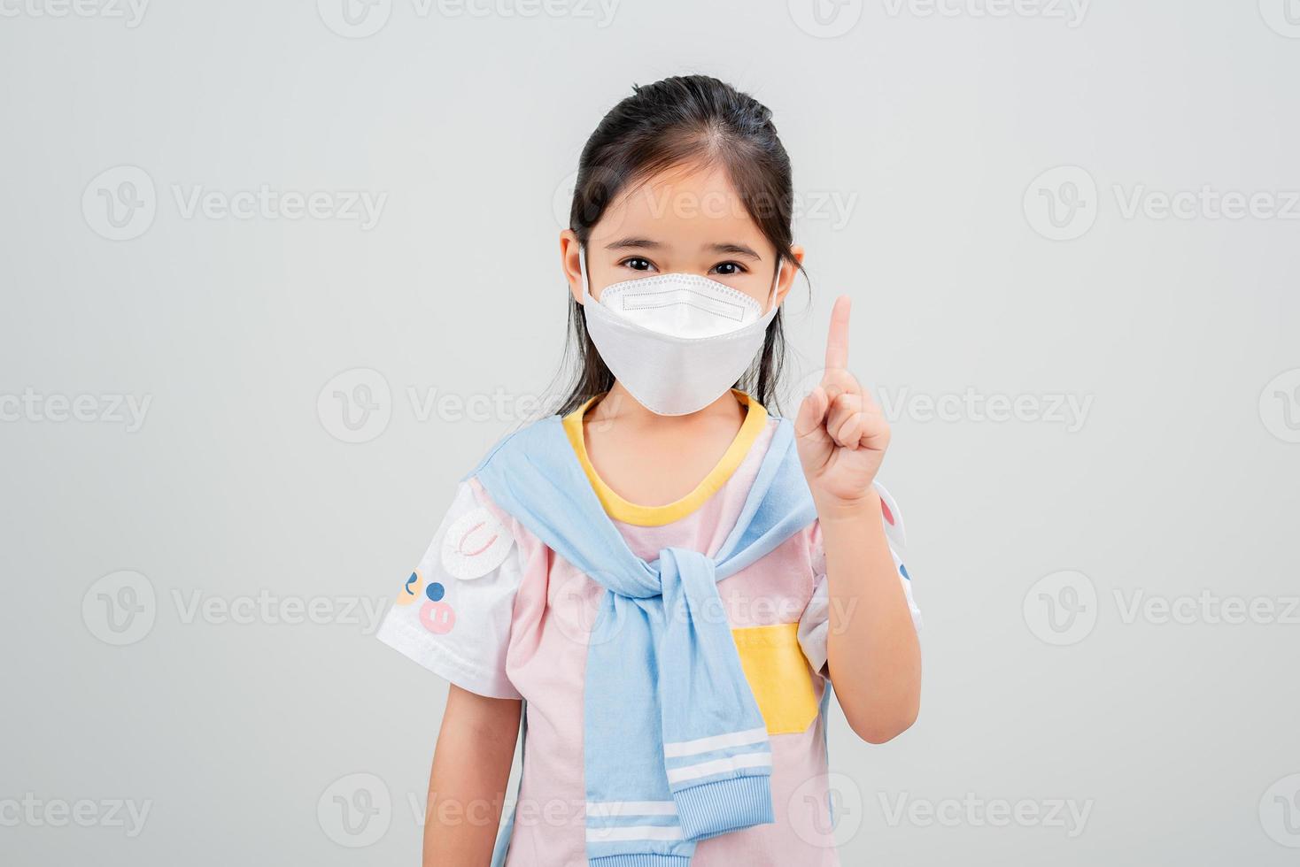 Asian little child girl wearing respirator mask to protect coronavirus outbreak and pointing hand to blank background, New virus Covid-19 photo