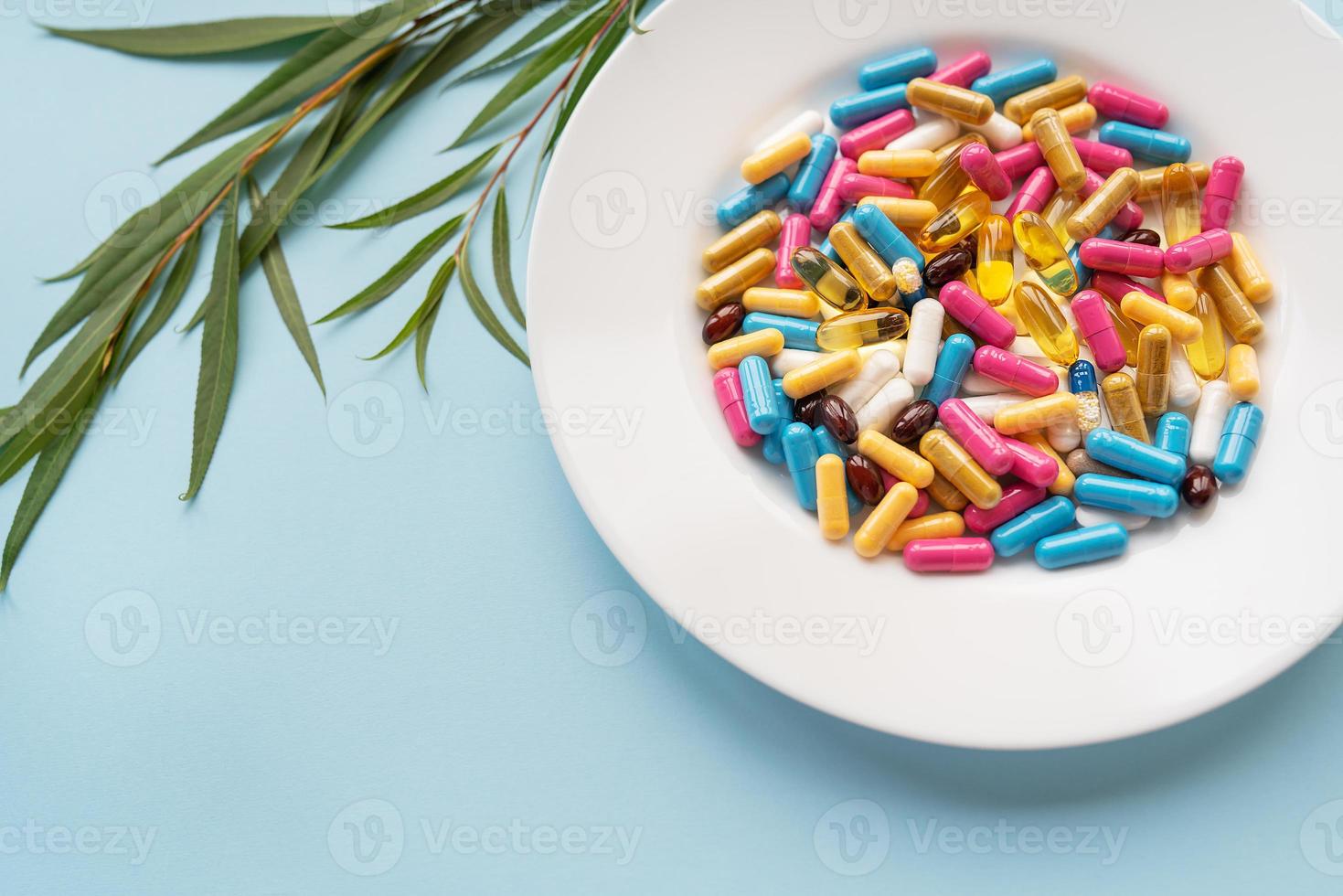 plato blanco con pastillas de suplementos nutricionales en diferentes colores brillantes. fondo azul, lugar para una inscripción. rama verde en el fondo. foto