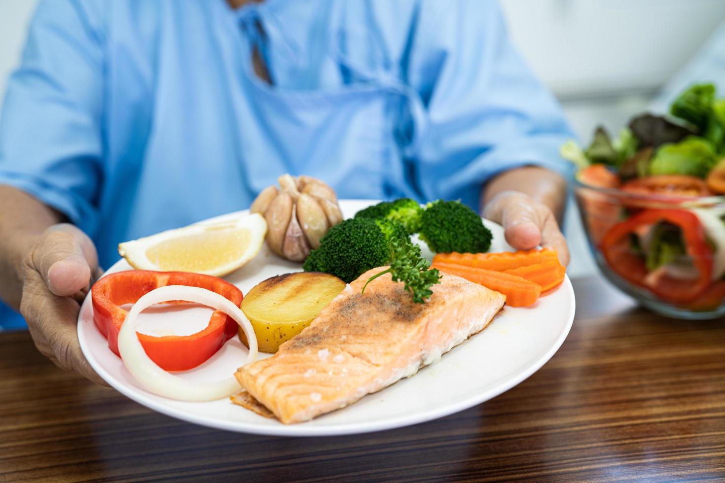 Asian senior or elderly old lady woman patient eating Salmon steak breakfast with vegetable healthy food while sitting and hungry on bed in hospital. photo