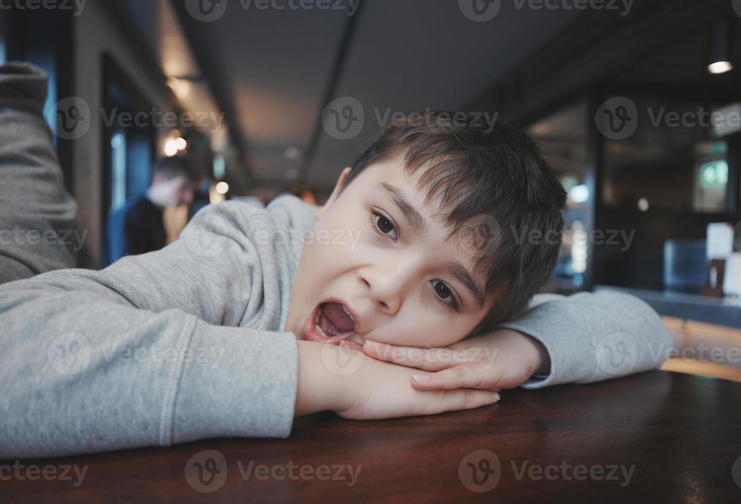 Kid looking out with bored face, Indoors Portrait preschool child laying head down on hand and yawning, Young boy sitting in Resturant or Cafe waiting for food photo