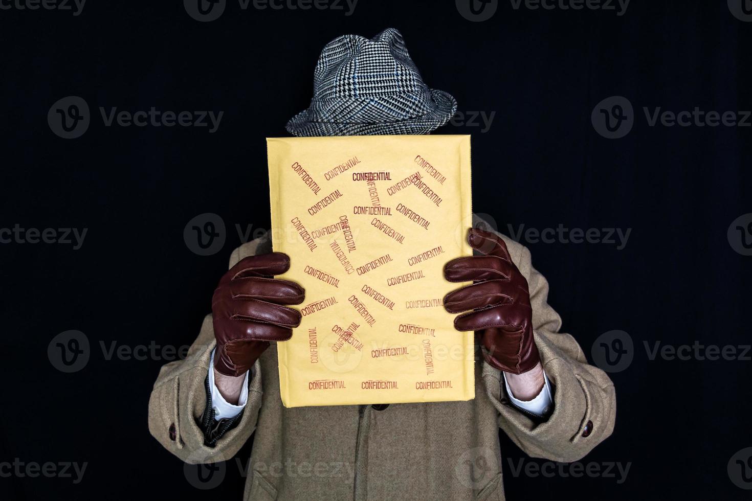 retrato de un hombre con abrigo y sombrero sosteniendo un sobre confidencial sobre su rostro con fondo negro. espía agente secreto de cine negro. robo de identidad y delincuencia. foto