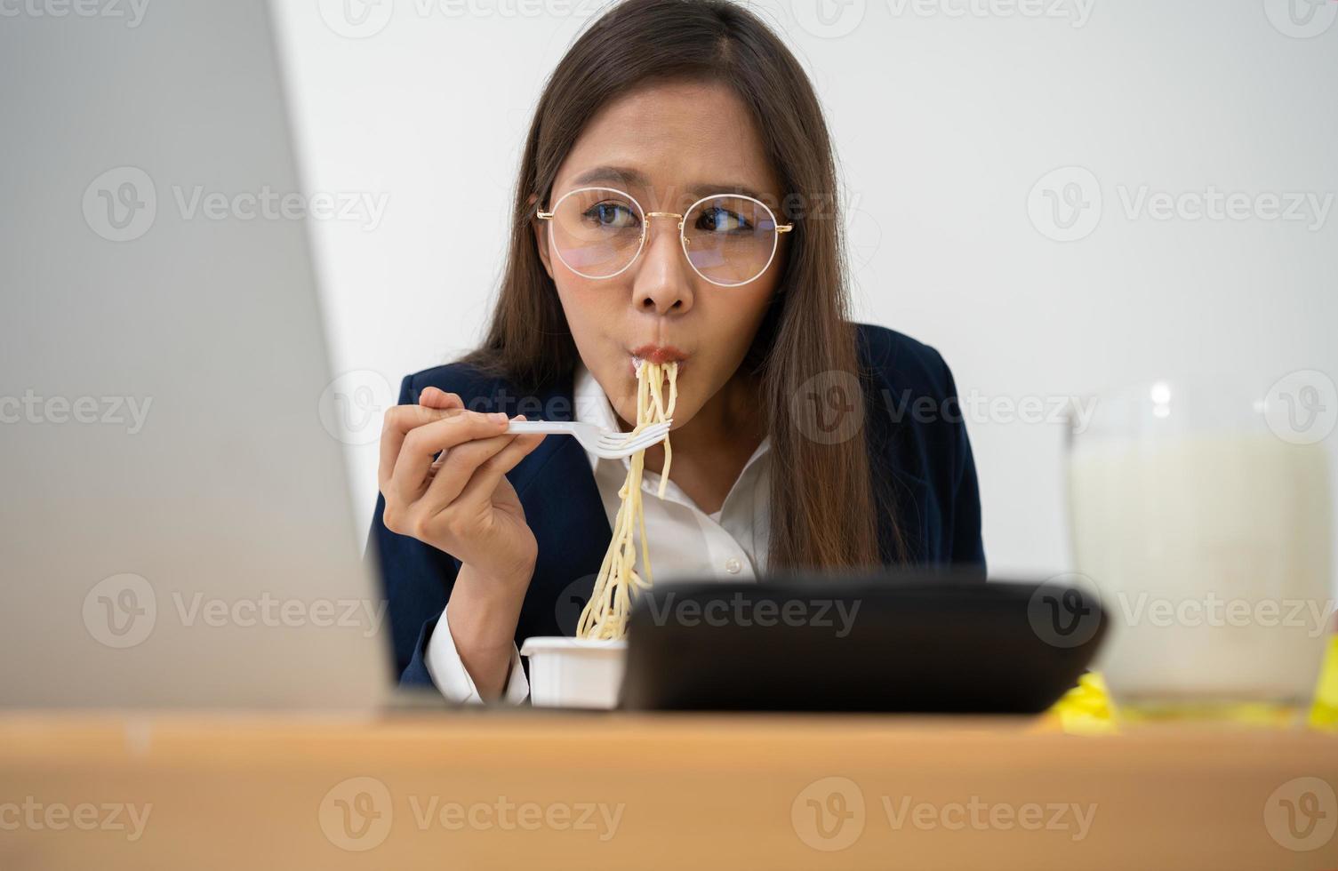 mujer de negocios ocupada y cansada comiendo espagueti para el almuerzo en la oficina y trabajando para entregar estados financieros a un jefe. con exceso de trabajo y poco saludable para comidas preparadas, concepto de agotamiento. foto