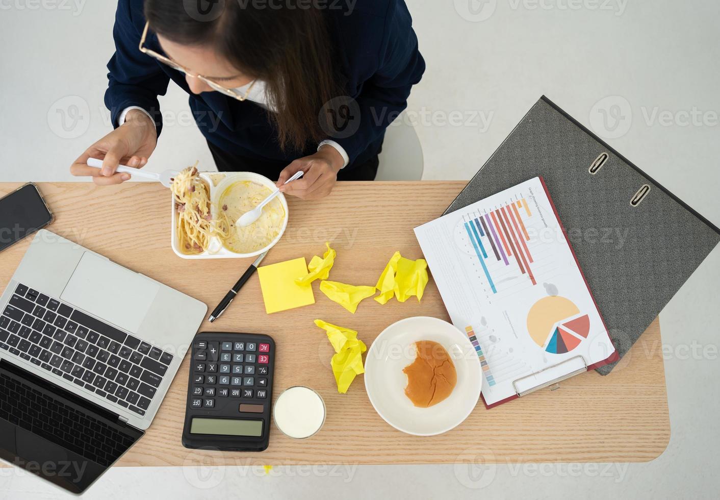 Top view of busy and tired businesswoman eating spaghetti for lunch at the Desk office and working to deliver financial statements to boss. Overworked and unhealthy for ready meals, burnout concept. photo