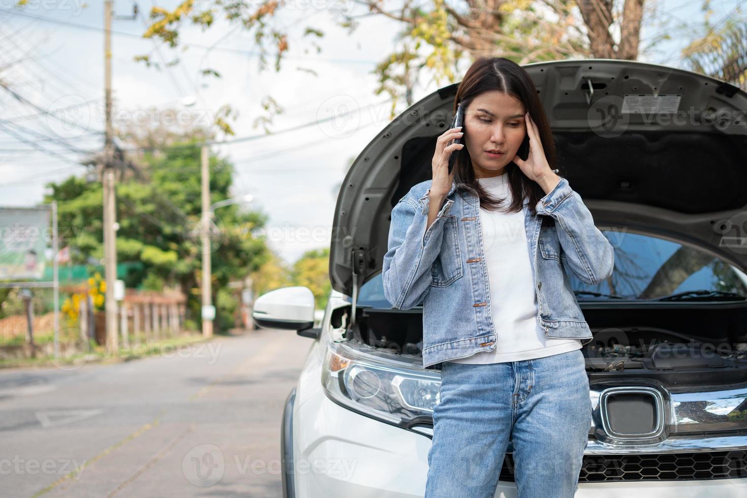 Angry Asian woman and using mobile phone calling for assistance after a car breakdown on street. Concept of vehicle engine problem or accident and emergency help from Professional mechanic photo