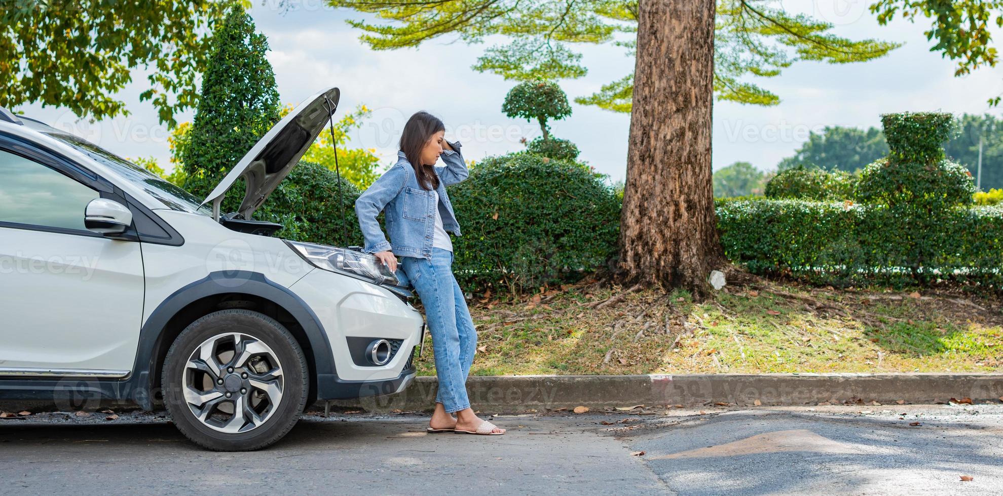 Angry Asian woman and using mobile phone calling for assistance after a car breakdown on street. Concept of vehicle engine problem or accident and emergency help from Professional mechanic photo