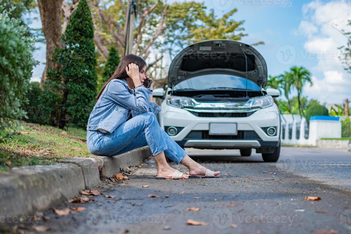 Asian woman sitting beside car after a car breakdown on street. Concept of vehicle engine problem or accident and emergency help from Professional mechanic photo
