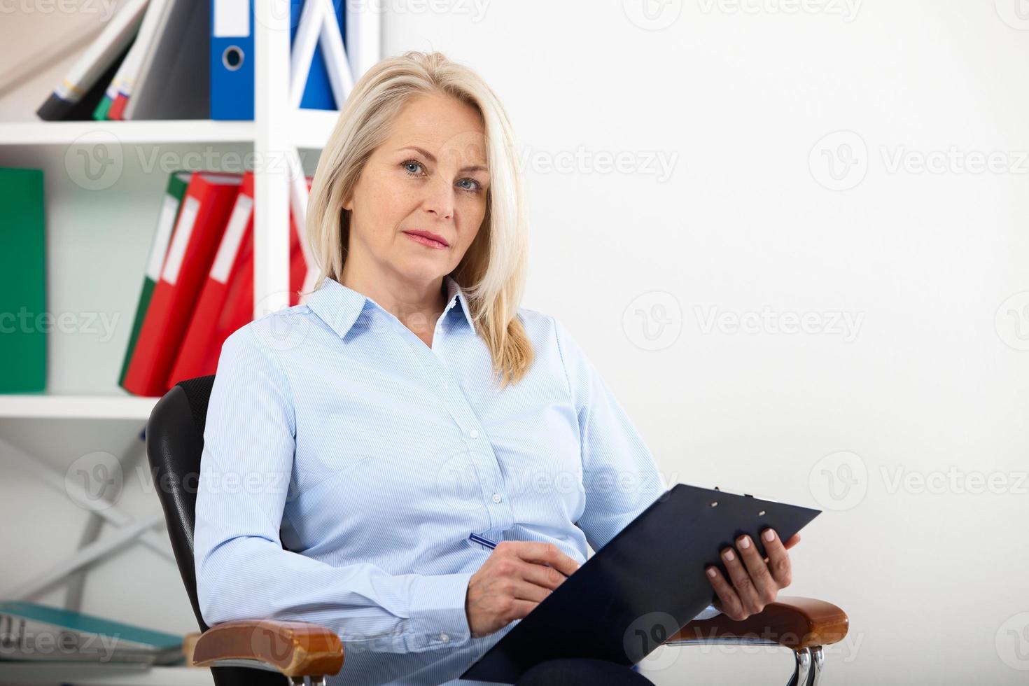 Business woman working in office with documents. Beautiful middle aged woman looking at camera with smile while siting in the office. photo