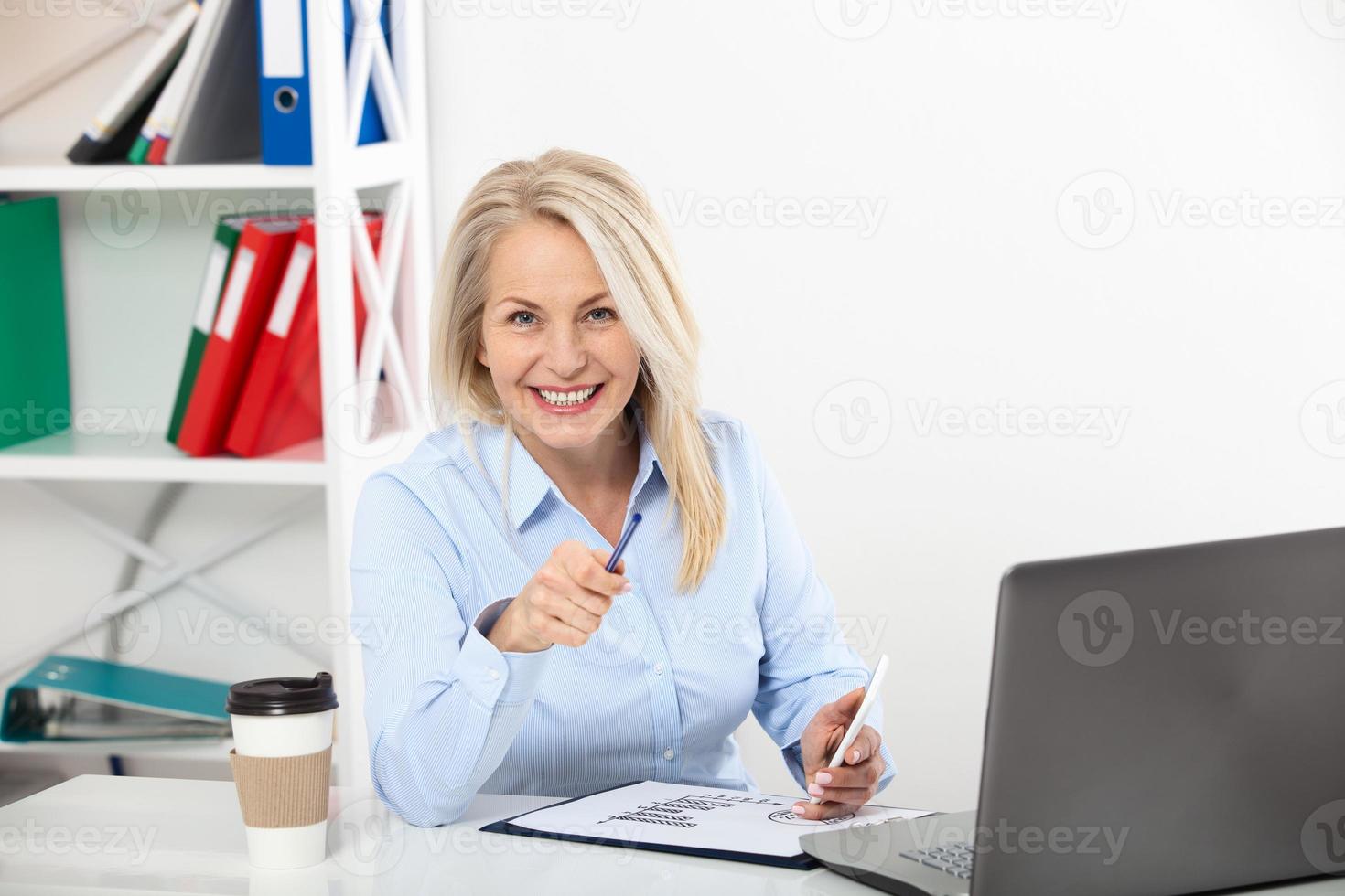 Business woman working in office with documents. Beautiful middle aged woman looking at camera with smile while siting in the office. photo