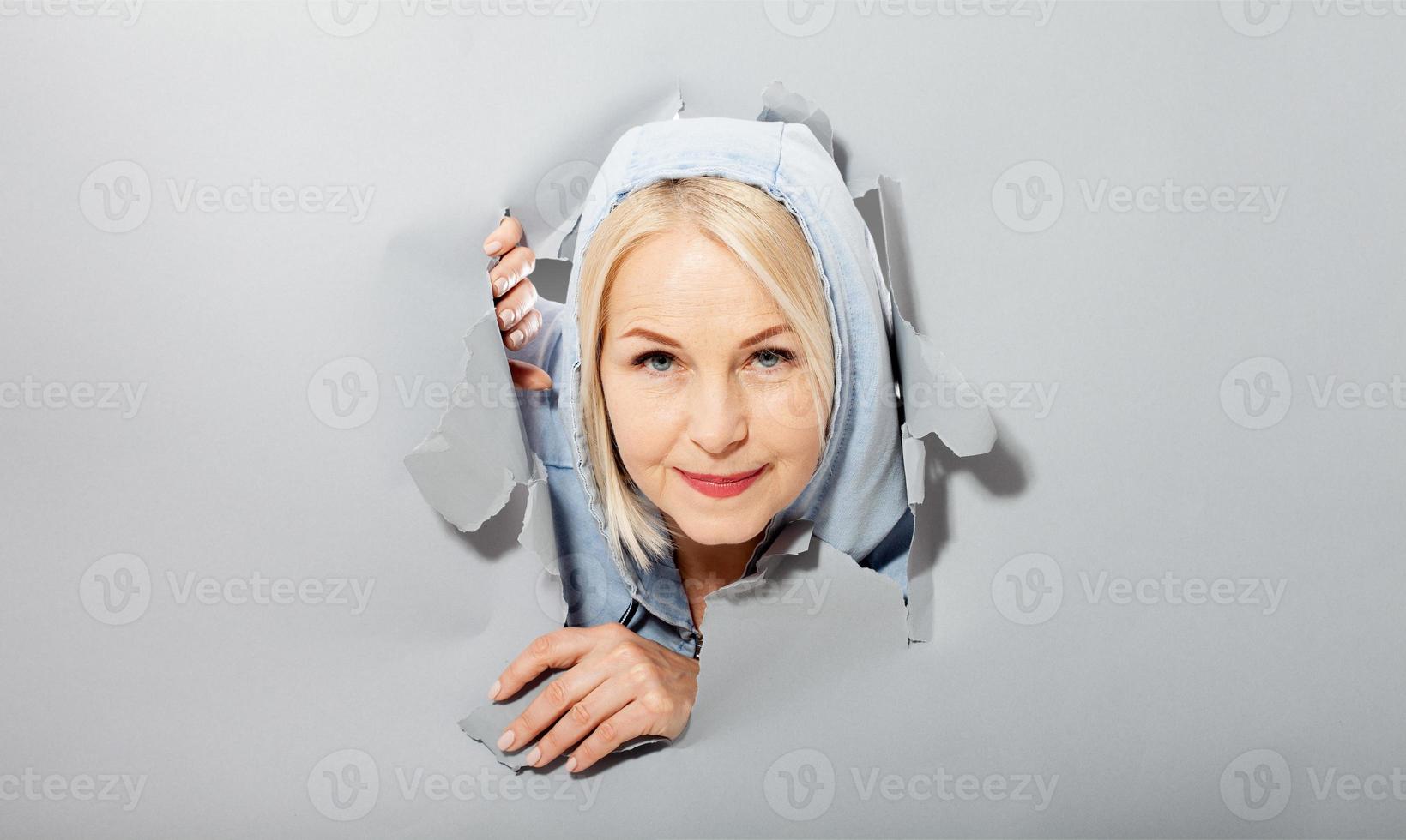 Charismatic joyful woman with blonde hairstyle, looking playfully, wears fashionable hoody, stands in torn paper hole, notices funny pleasant. Gray background. photo