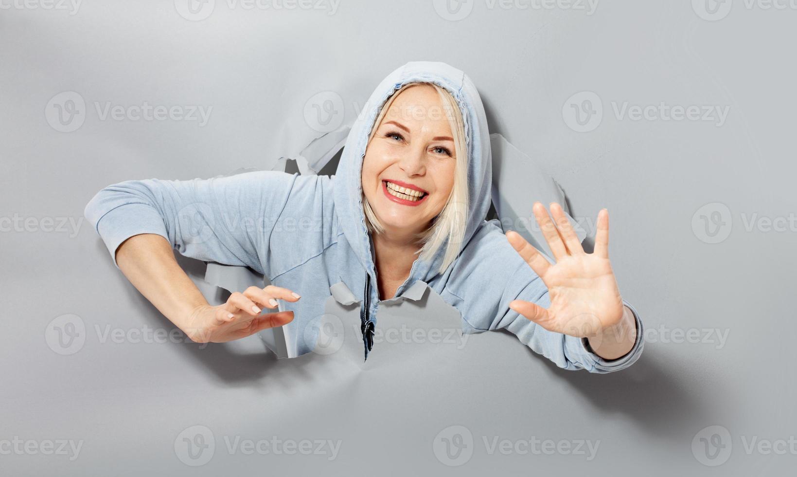 Charismatic joyful woman with blonde hairstyle, looking playfully, wears fashionable hoody, stands in torn paper hole, notices funny pleasant. Gray background. photo